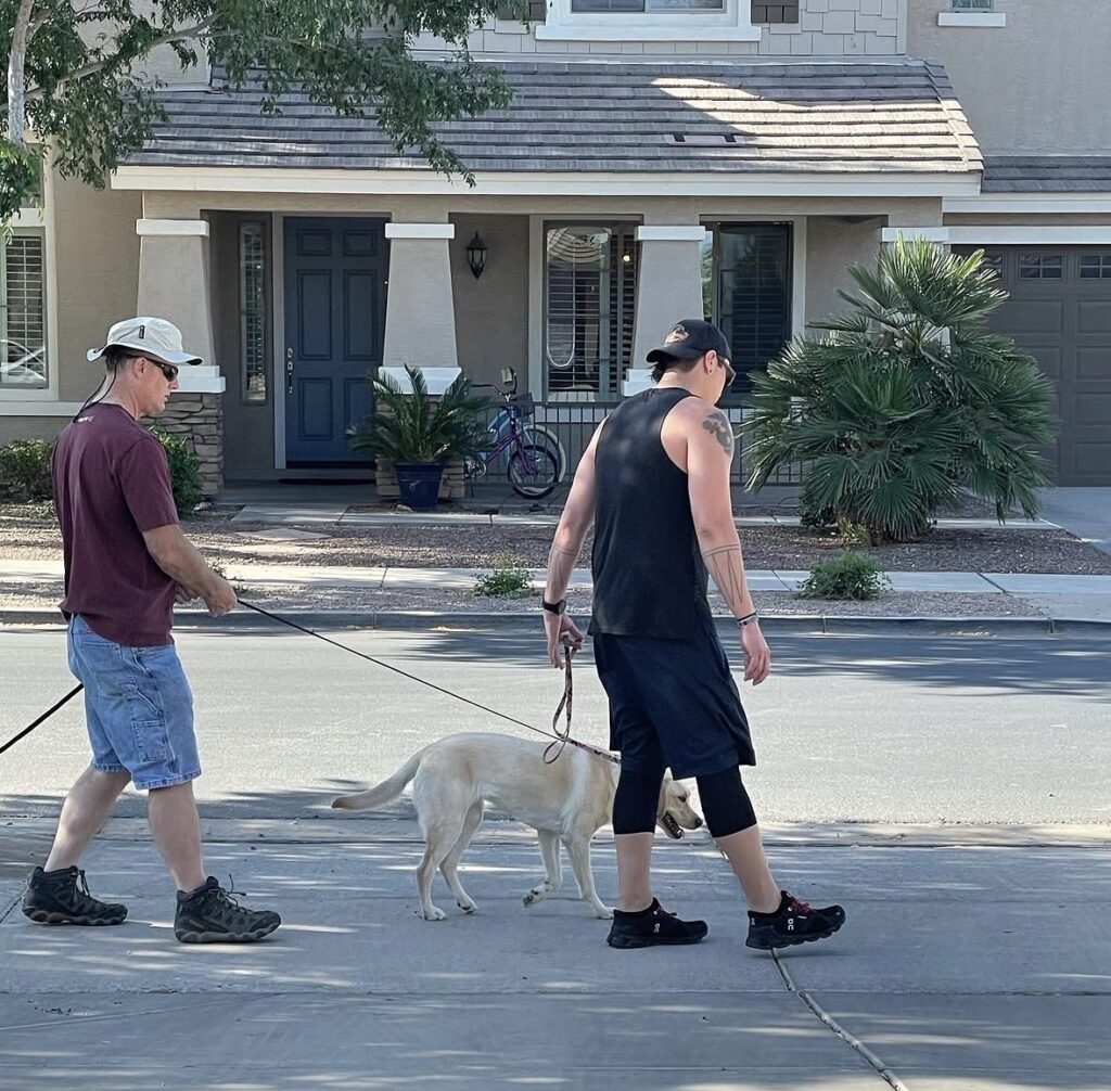 Sam Basso coaches a dog owner walking a light-colored dog on a leash along a residential sidewalk during private dog training in Gilbert, Arizona.